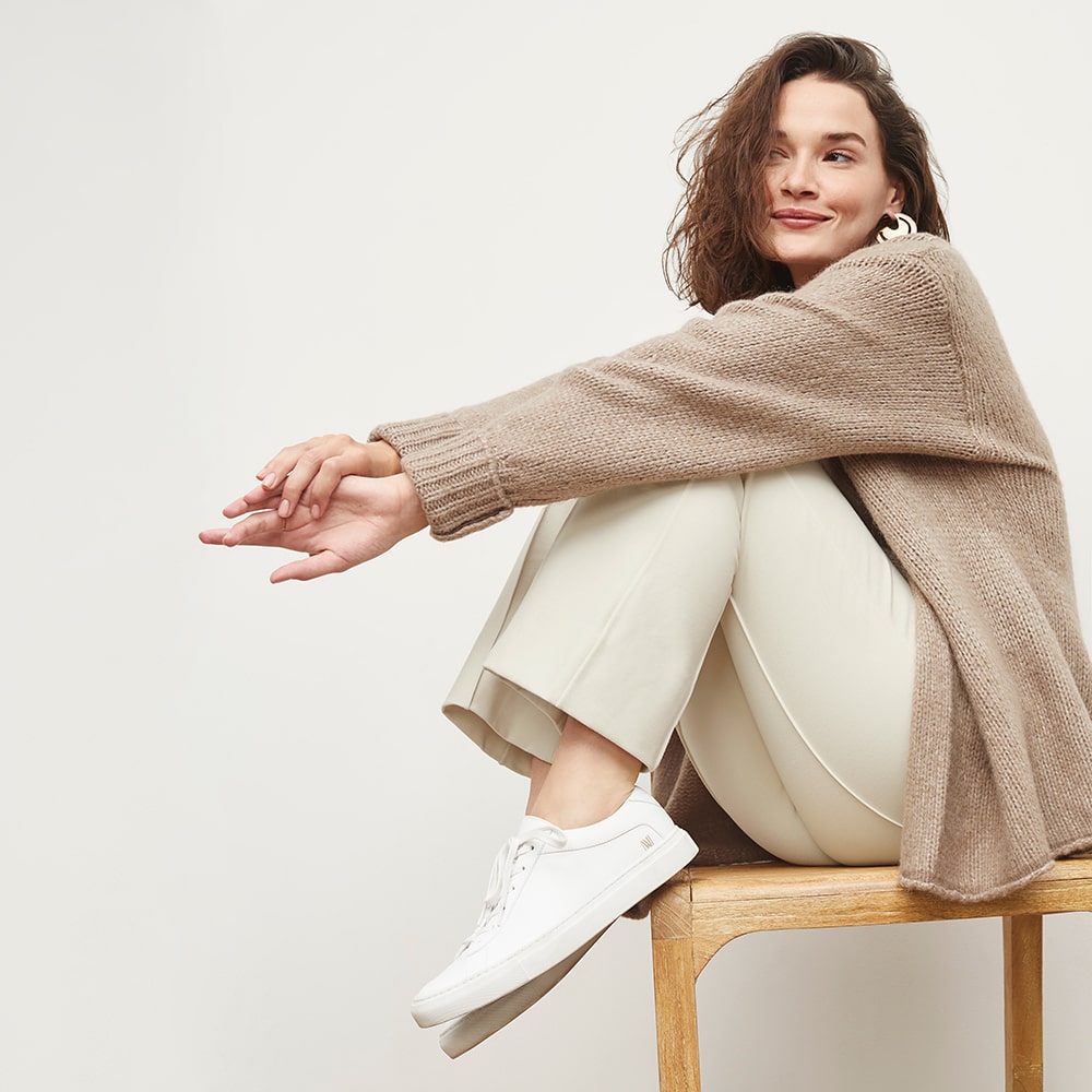 E-commerce imagery featuring a model in a cozy beige sweater and white sneakers, posed on a chair for a relaxed style.