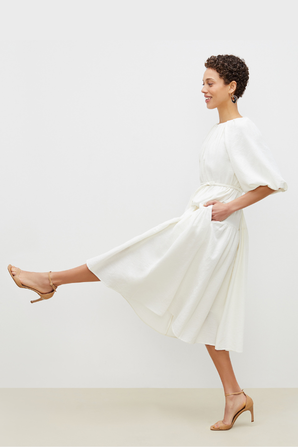 Woman in a flowing white MM LaFleur dress striking a playful pose, showcasing e-commerce photography for fashion and lifestyle brands